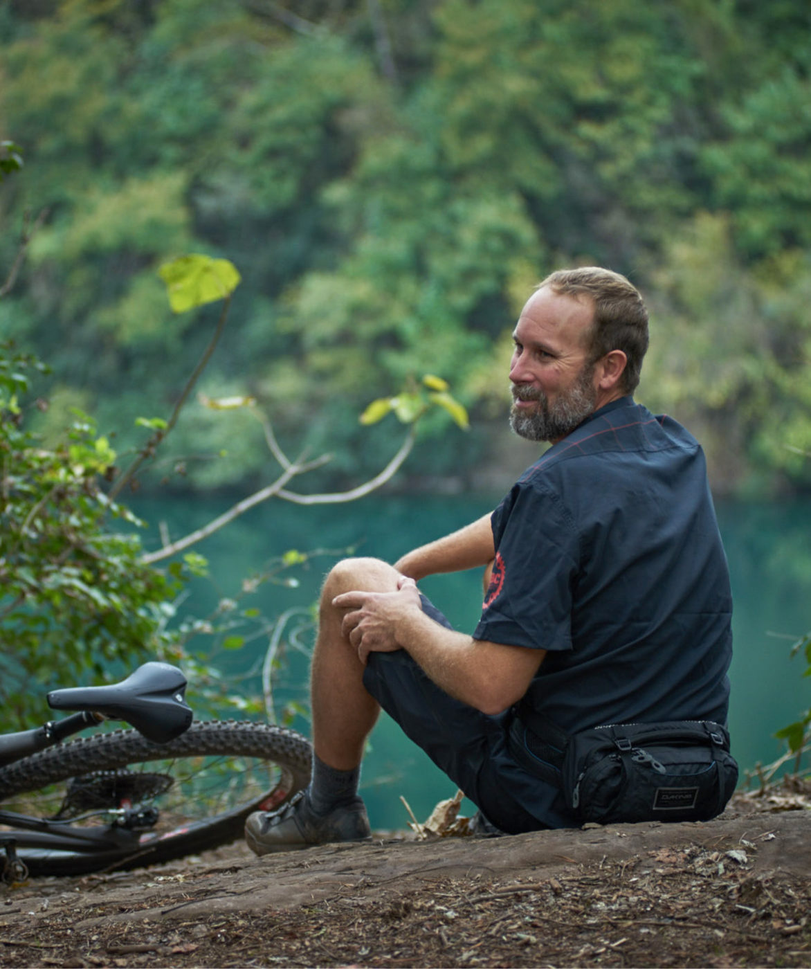 A man is seated on the ground next to his bicycle, overlooking the quarry lake and trees in the distance.