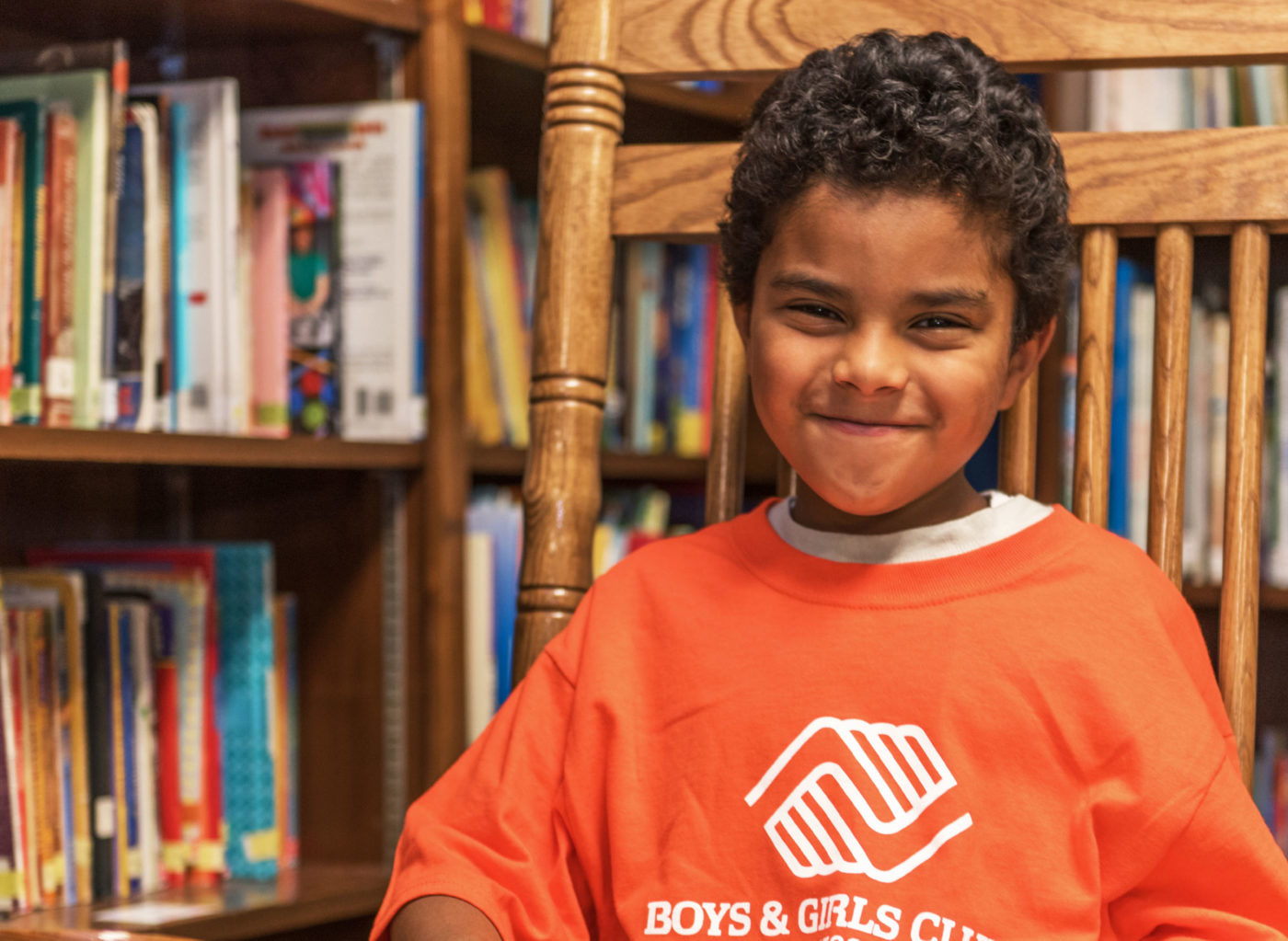 A young boy in an orange Boys & Girls Club t-shirt sits in a chair with a book in his lap.