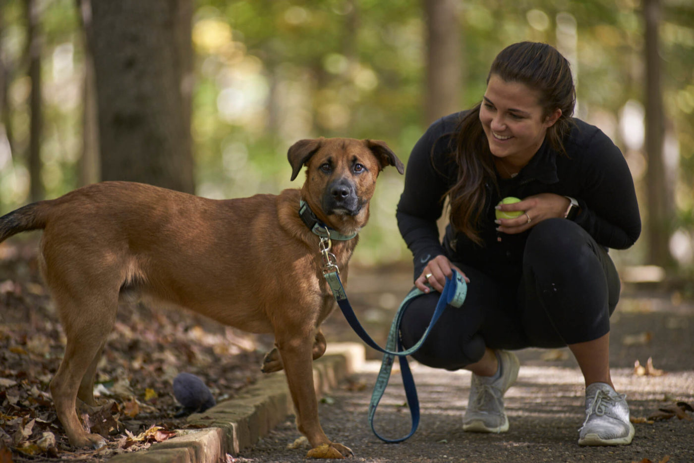 A woman dressed in black and holding a tennis ball smiles and crouches next to a brown dog on the trail.