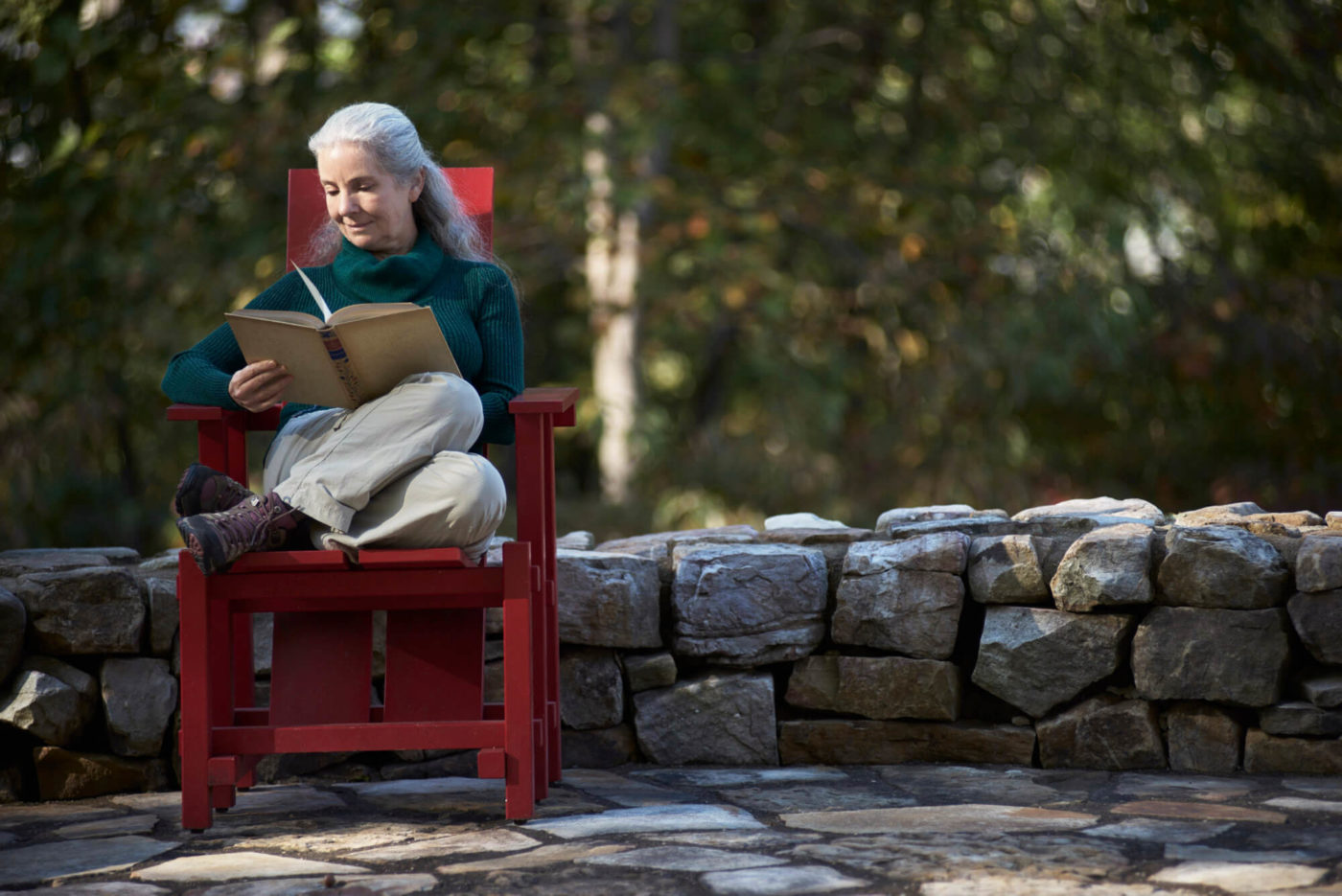 A woman with white hair sits and reads a book in a red Adirondack chair at High Ground Park.