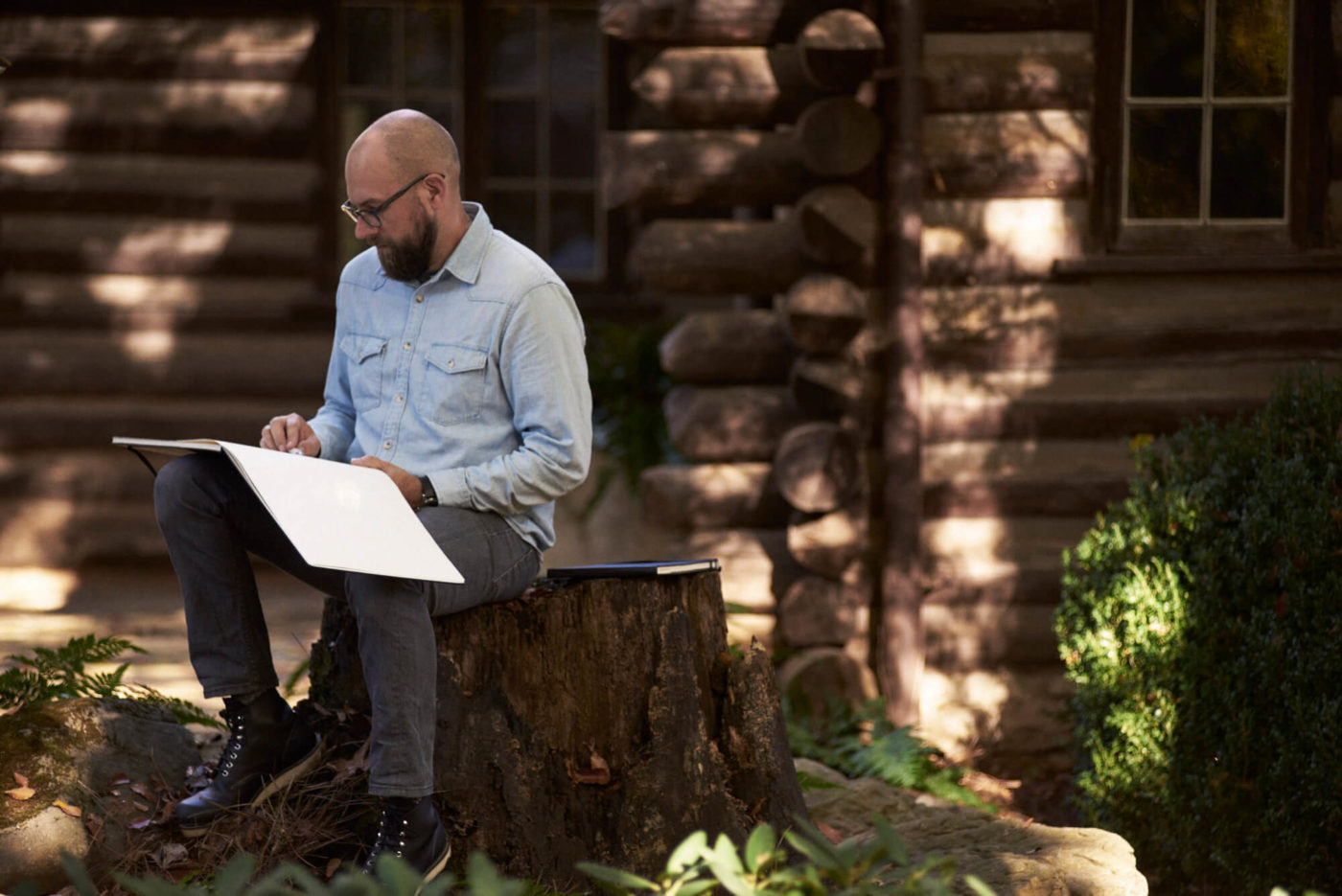 A man dressed in a blue button-down and gray slacks sits outside on a tree stump with a large sketchbook in his lap.