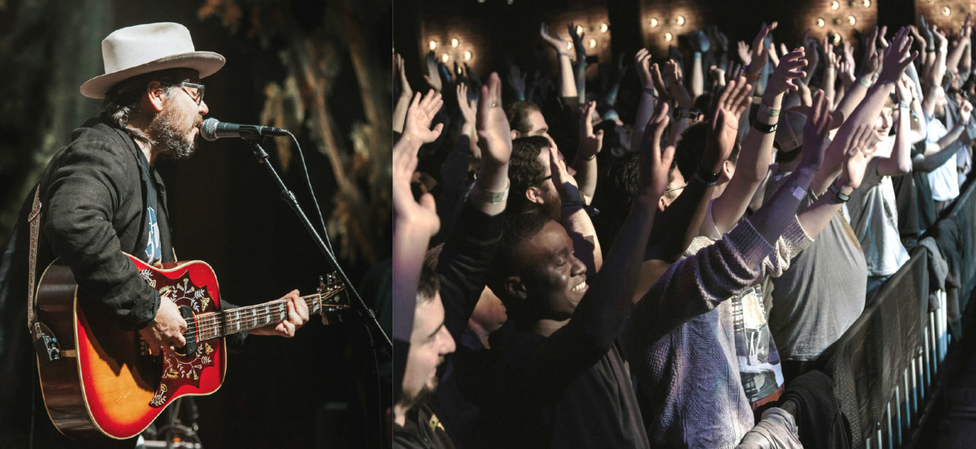 On the right: Jeff Tweedy of Wilco sings into a microphone and plays a red guitar. On the left: a concert audience raises their arms.