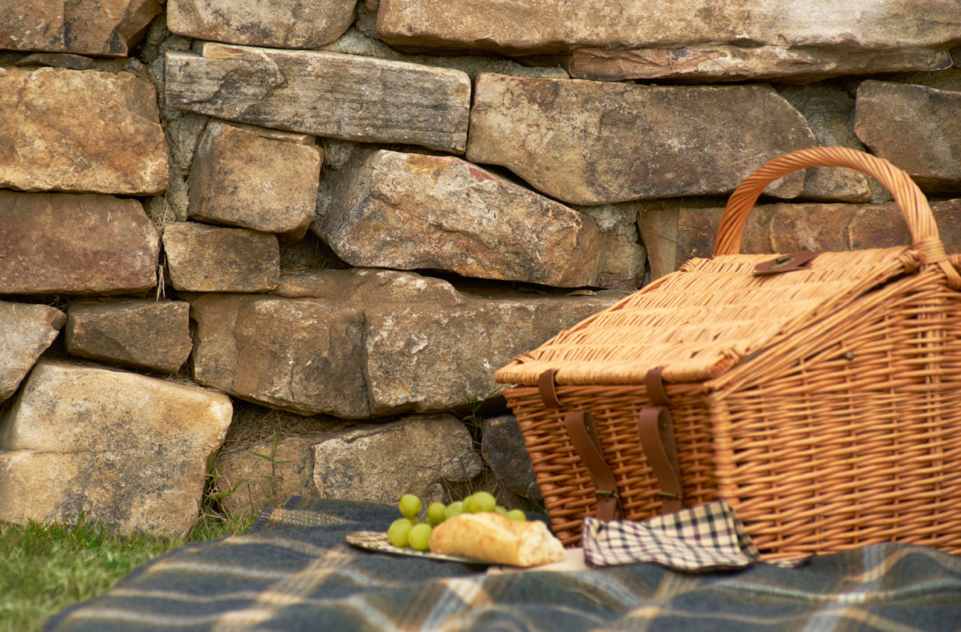 A picnic basket and a plate of food sits atop a plaid blanket next to a stone wall.