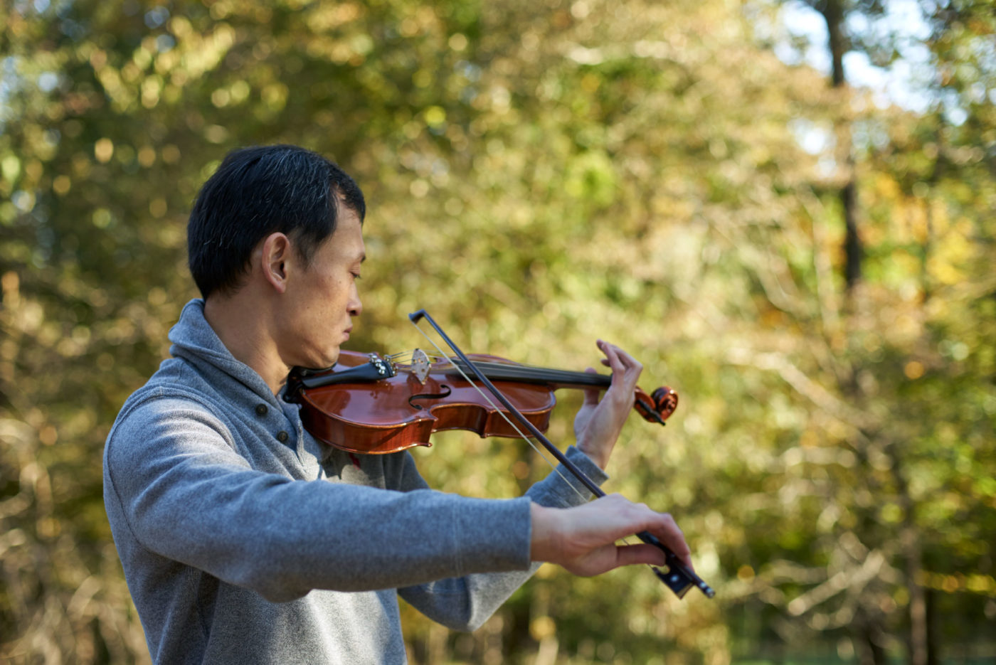 A man in a gray sweater plays the violin outside near the woods.