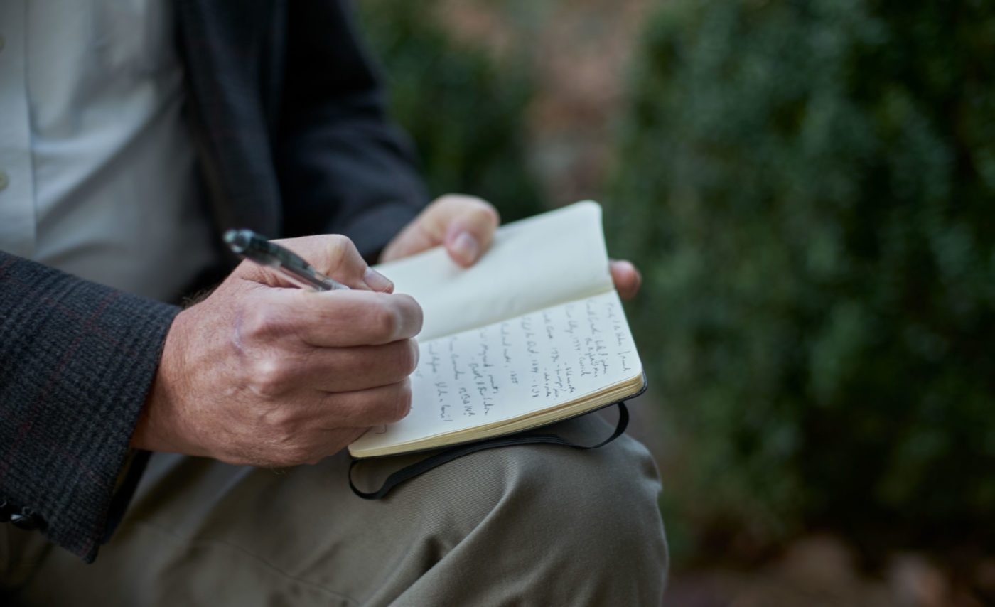 A close-up photo of a person seated outside, writing in a small notebook on their lap.