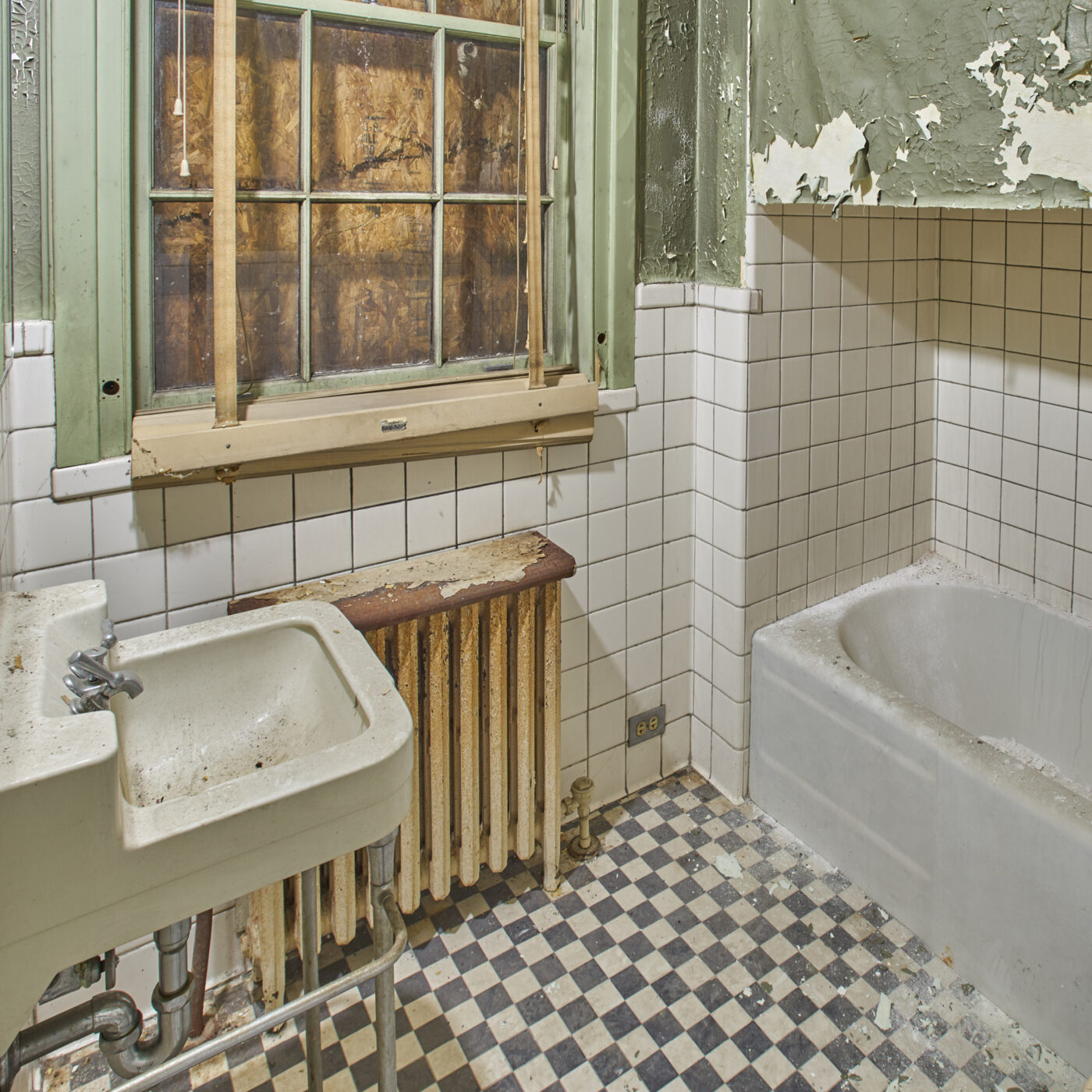 A photo of a bathroom sink and bathtub in pre-restoration condition. The window is boarded, the floor is tiled with black and white checkered tile, and the wallpaper is pale green.