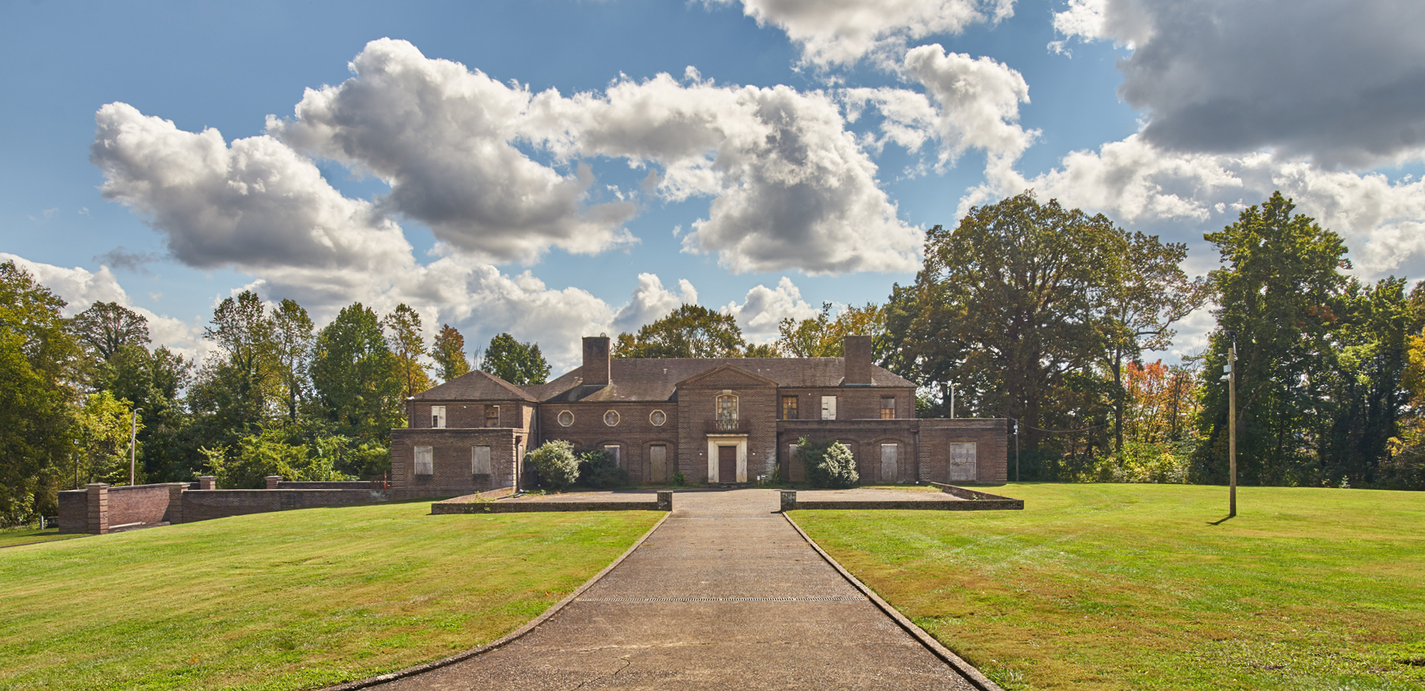 A large, brown brick house sits at the bottom of a grassy hill, with a blue sky full of cumulus clouds above.