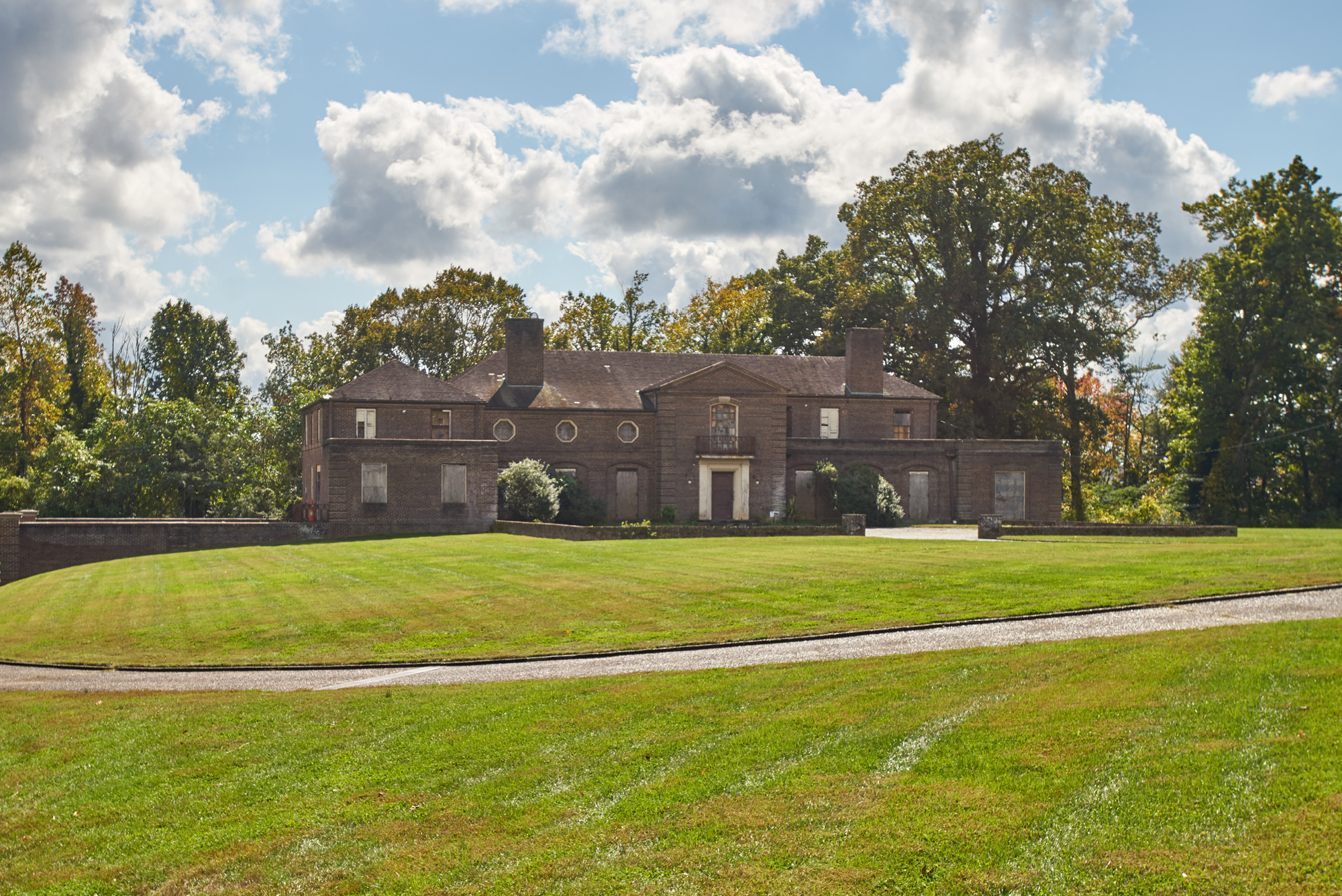 A large, brown brick house sits at the bottom of a grassy hill, with a blue sky full of cumulus clouds above.