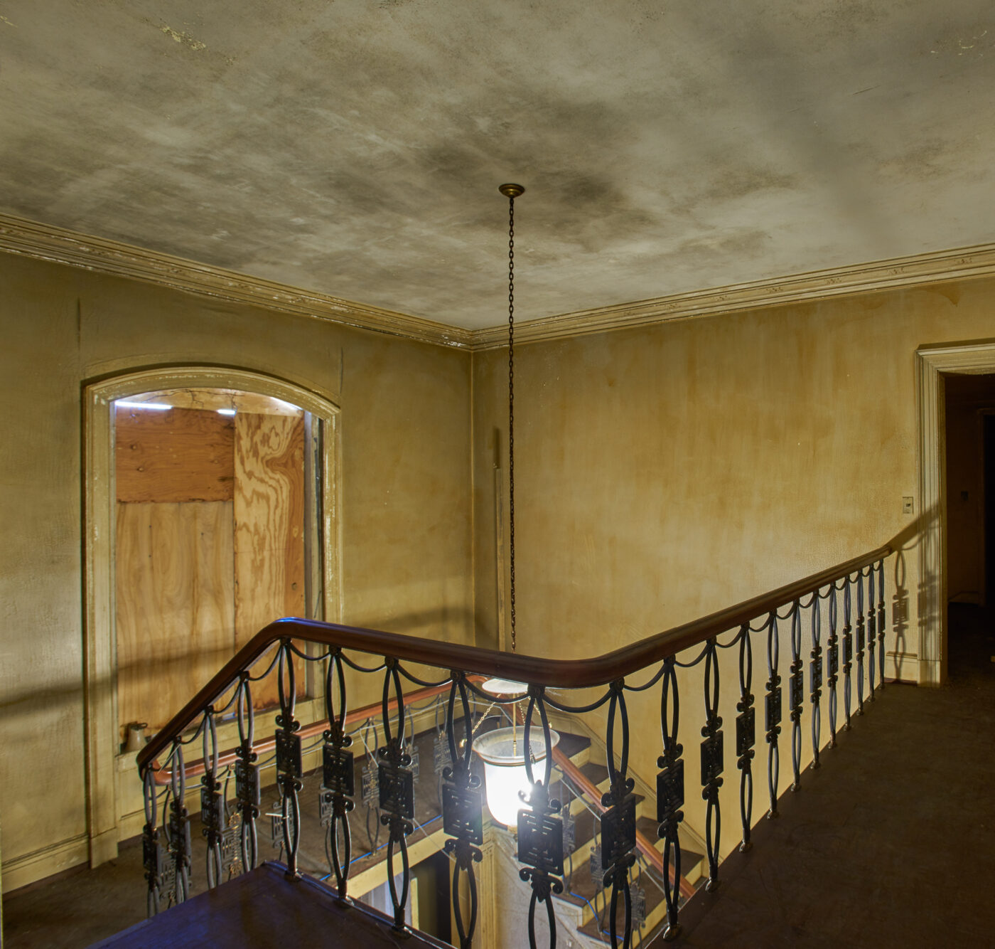 A photo of the second story landing, overlooking an ornate stair rail and a boarded-up second story window.