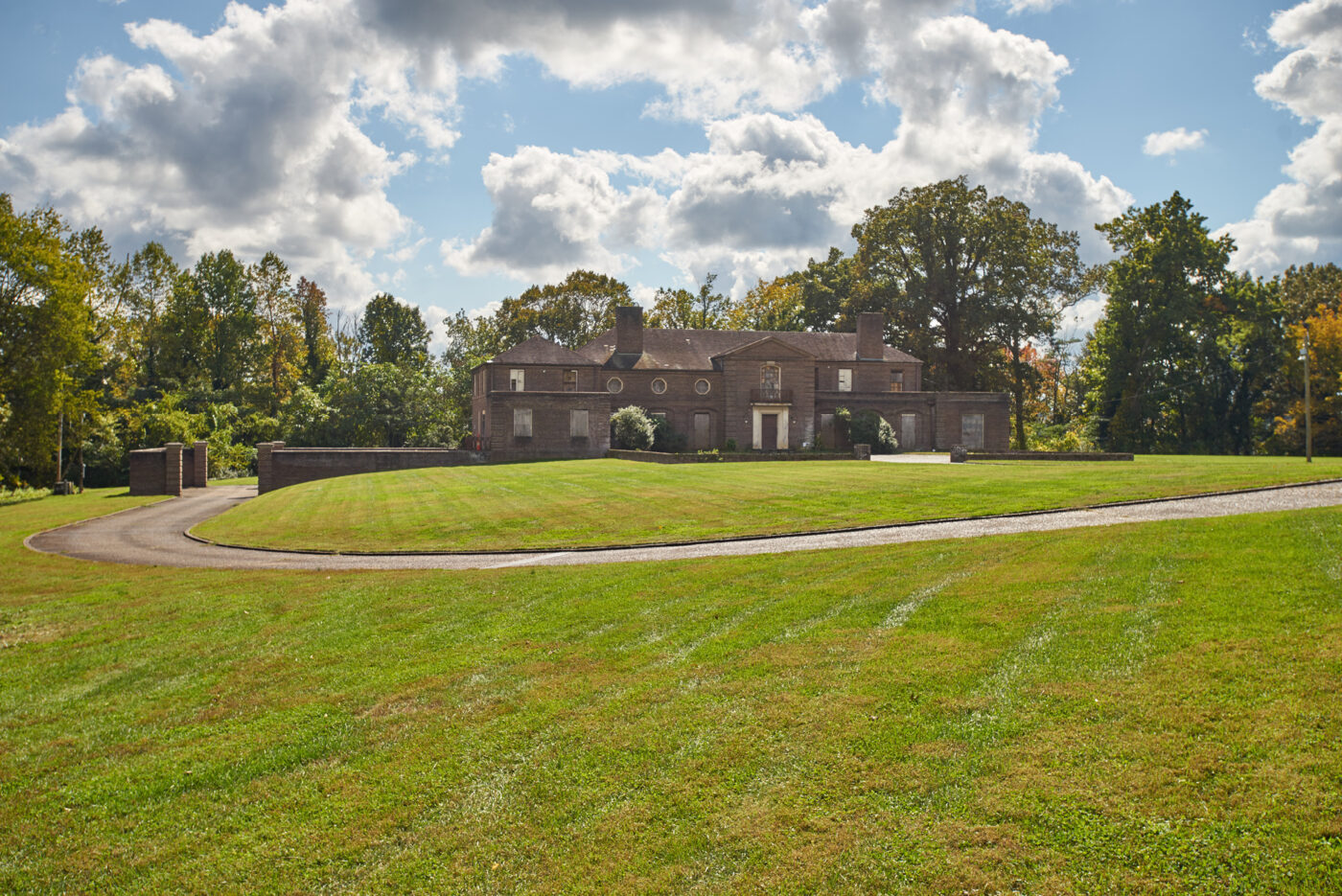 A large, brown brick house sits at the bottom of a grassy hill, with woods behind it and a blue sky full of cumulus clouds above.